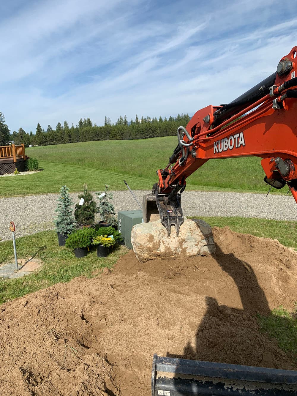 Kubota excavator digging in a yard with potted plants and trees nearby.