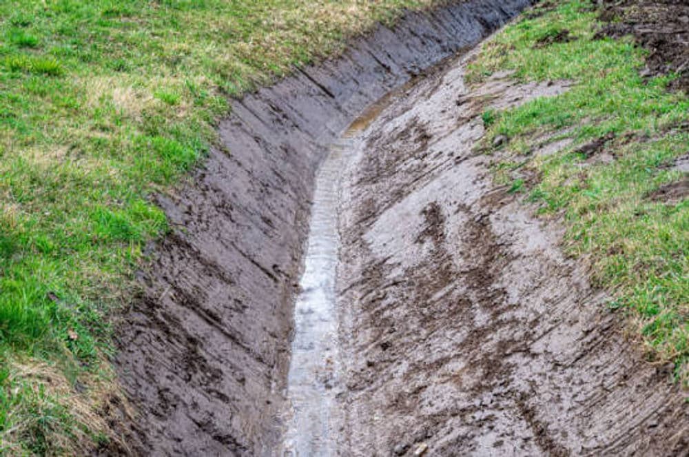 Water flowing through a muddy drainage ditch surrounded by green grass.